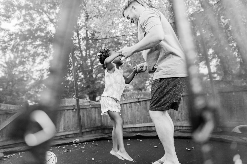 A dad jumps on a trampoline with his daughter as they hold hands during their lifestyle family photo session with Priscilla Baierlein.
