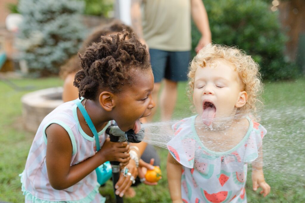 Two sisters drink from a water hose in a backyard garden in summer in Kentucky.