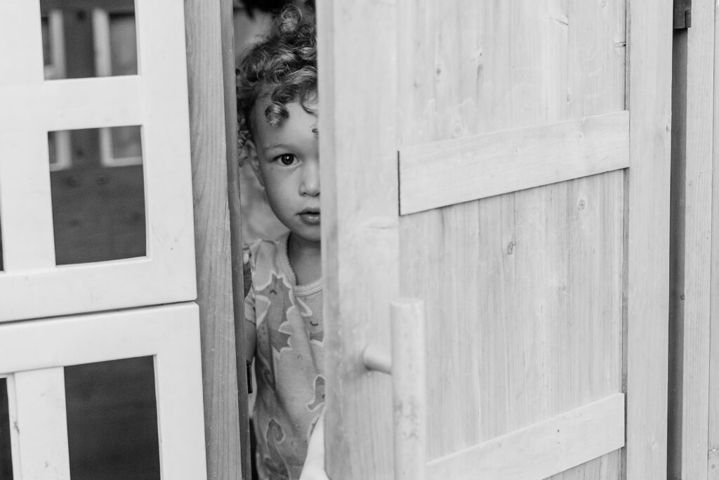 A black and white image of a little girl peaking out the door of her playhouse in her backyard during her family photo session in Lexington KY.