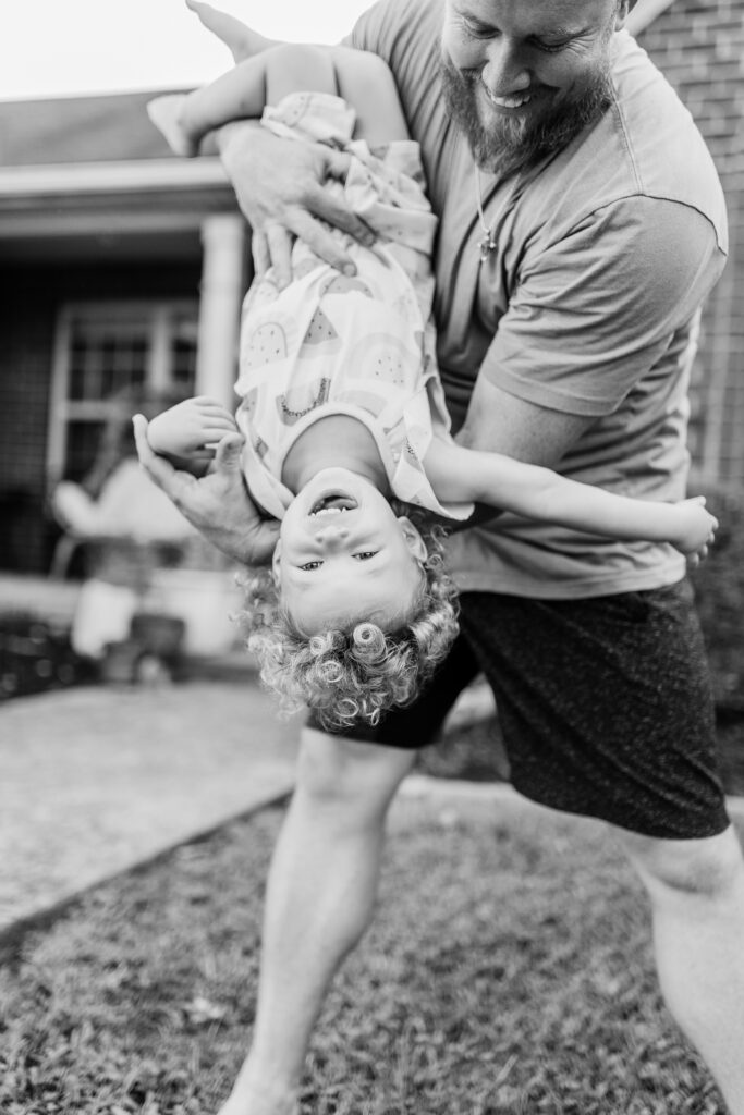 A black and white image of a dad playing with his little girl as he lifts her up and flips her upside down during a lifestyle family session in Lexington KY.