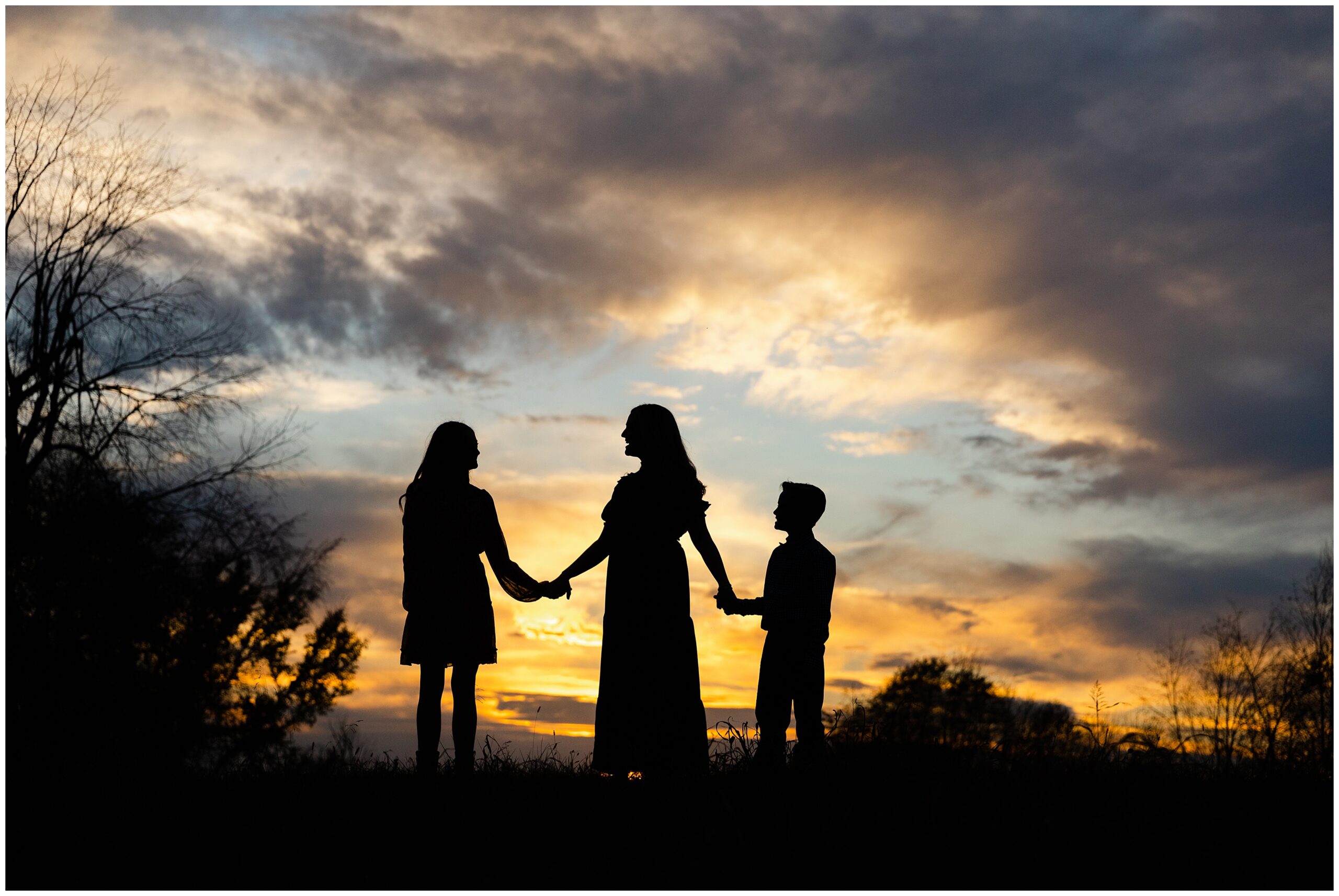 A sunset silhouette of a mother holding her son and daughters hands.