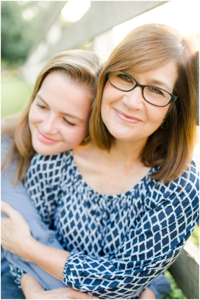 A mom looking gently at the camera while being hugged by her teenage daughter.